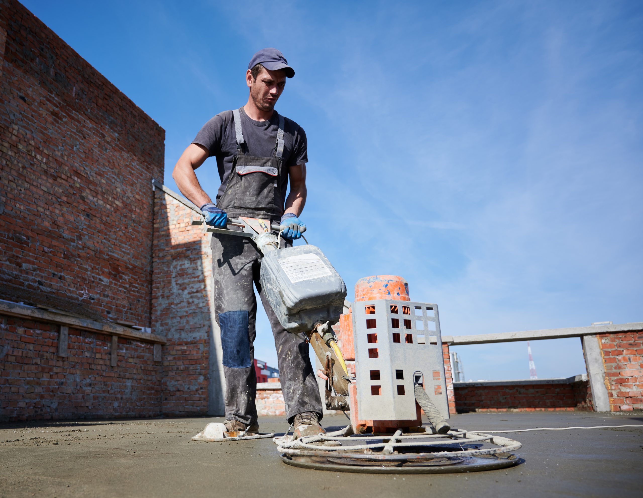 Full length of man laborer using troweling machine while screeding floor in new building under blue sky. Male worker finishing concrete surface with floor screed grinder machine at construction site.